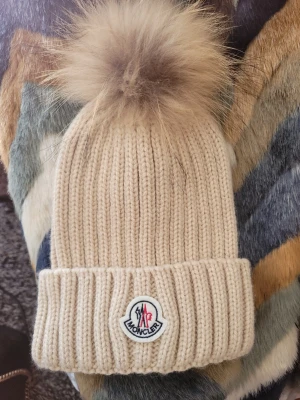 Beige Moncler mössa med pälsboll - Säljer en beige ribbstickad mössa från Moncler med stor fluffig pälsboll på toppen. Mössan har uppvikt kant och Moncler-logga framtill. Perfekt accessoar för att hålla stilen när det är kallt ute. Endast testad. Ny pris 3047kr 