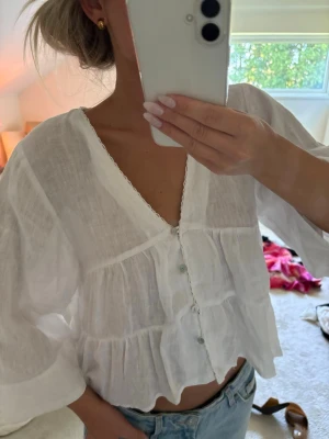 Vit luftig blus med knappar - Säljer en vit, luftig blus med v-ringning och vida ärmar. Blusen har dekorativa knappar framtill och är gjord i ett tunt, lätt material som känns som bomull eller linne. Perfekt till jeans eller kjol för en avslappnad look.