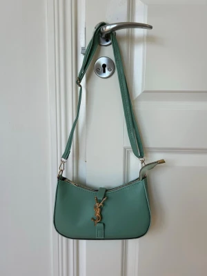 Grön axelremsväska med gulddetaljer - Snygg ljusgrön handväska i slätt konstläder med justerbar axelrem och guldfärgad dragkedja. Väskan har en elegant YSL-liknande metallogo framtill och minimalistisk design. Perfekt storlek för det viktigaste och passar till många outfits.