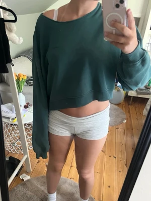 Grön croppad sweatshirt - Mörkgrön croppad sweatshirt med bred halsringning och långa ärmar. Tröjan har en avslappnad passform och ribbade muddar vid ärmslut och nederkant. Perfekt för en chill och trendig look.