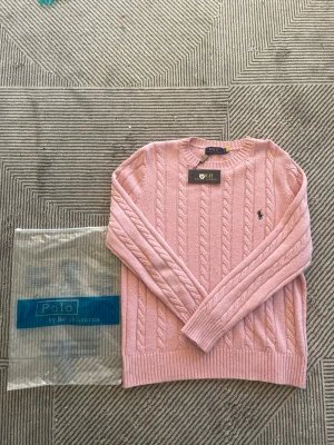 Rosa kabelstickad tröja Polo Ralph Lauren - Supersnygg rosa kabelstickad tröja från Polo Ralph Lauren. Klassisk rund halsringning, långa ärmar och ribbade muddar. Ikoniska loggan broderad på bröstet. Perfekt för dig som vill ha en stilren och trendig look.