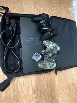 PlayStation 2 med tillbehör - PlayStation 2 konsol i svart med två handkontroller (en svart och en genomskinlig), minneskort och kablar. Allt ligger i en praktisk svart väska. Perfekt för dig som vill spela klassiska PS2-spel och återuppleva nostalgin.