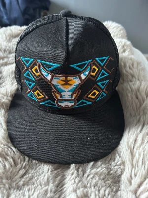 Svart keps med broderad tjur - Säljer en svart keps med platt skärm och mesh på baksidan. Framtill finns ett stort broderi av ett tjurhuvud i turkos, brunt och vitt, omgiven av geometriska mönster i turkos, gult och brunt. Kepsen har en modern streetwear-vibe och justerbar snapback.