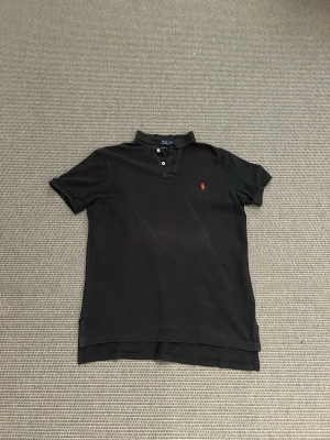 Ralph lauren piké - Klassisk svart pikétröja från Ralph Lauren med röd broderad logga på bröstet. Tröjan har korta ärmar, krage och knappar framtill. Tillverkad i bomull och har en snygg, normal passform. Perfekt för en clean och stilren look.