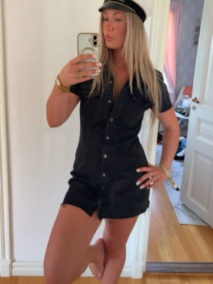 Svart kort jeansklänning - Säljer en svart kort jeansklänning med knappar framtill och korta ärmar. Klänningen har en klassisk skjortkrage och en figurnära passform som ger en snygg siluett. Perfekt för dig som gillar en enkel men trendig look.  Är storlek M men passar även S