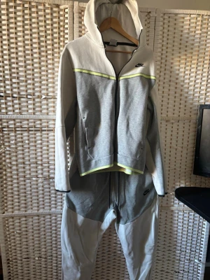 Nike grå och vit loungewear set NIKE TECH - Nike loungewear set med hoodie och byxor i grått och vitt med ljusgula detaljer. Setet har dragkedja framtill på hoodien och fickor på både överdel och byxa. Tillverkad i mjukt bomullsmaterial för maximal komfort. Perfekt för chill dagar hemma.