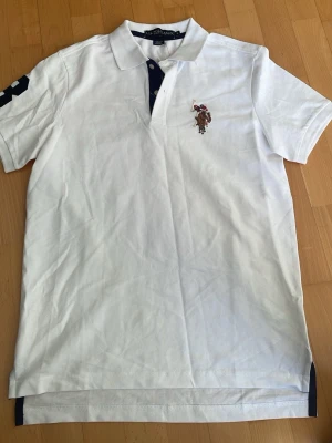 Vit pikétröja från U.S. Polo Assn. - Klassisk vit pikétröja från U.S. Polo Assn. med broderad logga på bröstet och kontrastfärgade detaljer vid knapparna. Tröjan har korta ärmar och krage, samt en siffra på ena ärmen för en sportig look.
