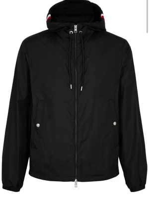 Svart moncler windbreaker  - Stilren svart vindjacka med huva och dragsko, hel dragkedja framtill och två fickor med tryckknappar. Jackan har en diskret röd och vit rand på huvan samt en patch på ärmen. Perfekt för blåsiga dagar och enkel att matcha.