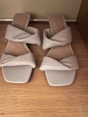 Beige slip-in sandaler med klack - Snygga beige sandaler med fyrkantig tå och vadderade, korslagda remmar. Slip-in modell med låg klack och mjuk innersula för extra komfort. Perfekta för dig som gillar trendiga och stilrena skor till sommaren.