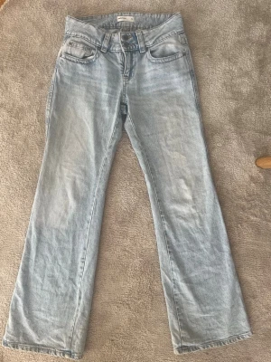 Ljusblå raka jeans med dubbla knappar - Säljer ett par ljusblå jeans med raka ben, lite utsvängda i ändarna och dubbla knappar i midjan. Jeansen har klassisk femficksdesign och är tillverkade i denim. Perfekta för en avslappnad och trendig look.