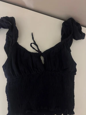Mörkblå croppad topp från Bershka - Säljer en svart croppad topp från Bershka med puffiga axelband och smockad midja. Toppen har en söt knytning framtill och volangkant nedtill. Perfekt för dig som gillar trendiga och feminina detaljer.