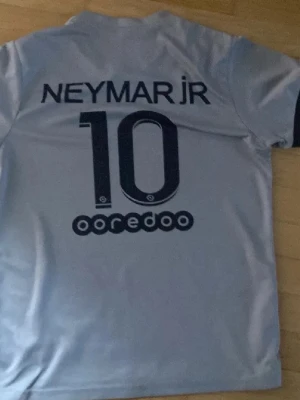 Grå Neymar Jr fotbollströja - Säljer en grå fotbollströja med Neymar Jr och nummer 10 tryckt på ryggen. Tröjan har korta ärmar och svart tryck, samt Ooredoo-logga under numret. Perfekt för dig som gillar fotboll och vill bära din favoritspelares namn.
