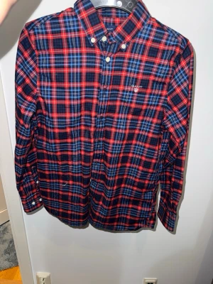 Gant skjorta - Snygg rutig skjorta från Gant i rött, blått och vitt. Skjortan har klassisk krage med knappar, lång ärm och broderad logga på bröstet. Perfekt för dig som gillar en tidlös och stilren look. Skjortan är nästan helt oanvänd och storleken syns på sista bilden. Ny pris ca 800 kom hjärna med prisförlag!