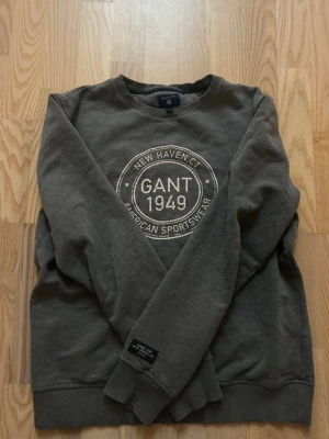 Grå sweatshirt från GANT - Mysig grå sweatshirt från GANT med rund halsringning och tryck på bröstet där det står 'GANT 1949 New Haven CT American Sportswear'. Tröjan har ribbade muddar vid ärmslut och nederkant samt en klassisk relaxed fit.