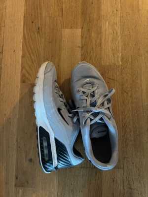 Grå Nike Air Max sneakers - Snygga grå Nike Air Max sneakers med vit sula och svart detalj vid hälen. Skorna har mesh-ovandel för bra andningsförmåga, klassisk snörning och synlig Air Max-dämpning i sulan. Perfekta för dig som gillar sportig och stilren look.