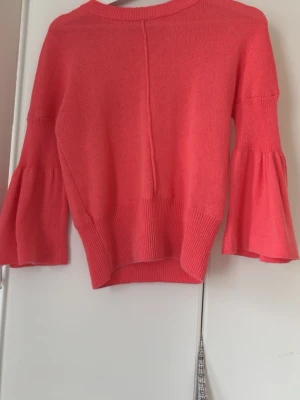 Korallfärgad stickad tröja med volangärm - Trendig stickad tröja i korall med bred ribbad kant och vida volangärmar. Tröjan har en rak passform och en dekorativ söm mitt bak. Perfekt för dig som vill ha en färgklick i garderoben.