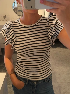Randig topp med volangärmar - Säljer en ribbad topp med svarta och vita ränder och volangiga korta ärmar. Toppen har en rund halsringning och sitter tight. 