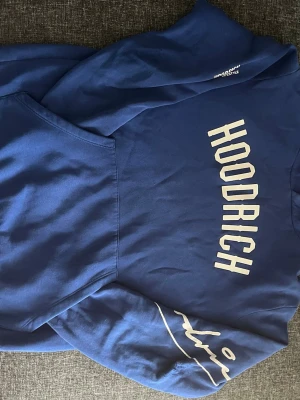 Blå HOODRICH hoodie med tryck - Snygg blå hoodie från HOODRICH med stort vitt tryck på bröstet och coolt grafiskt tryck på ena ärmen. Klassisk huva och känguruficka framtill. Perfekt för en streetwear-look och riktigt skön att bära till jeans eller joggers.