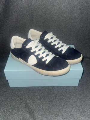 Mörkblå Phillipe model  med vita detaljer - Snygga mörkblå sneakers från Philippe Model med vita skosnören, vit sula och vita detaljer på häl och sida. Ovandelen är i mocka och skon har en klassisk låg profil som passar till det mesta. Perfekta för dig som gillar stilrena och trendiga skor.