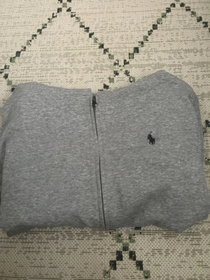 Grå zip hoodie från Polo Ralph Lauren - Snygg gråmelerad hoodie med dragkedja från Polo Ralph Lauren. Klassisk design med huva och två fickor framtill. Liten mörkblå logga broderad på bröstet. Perfekt för en chill och avslappnad stil.