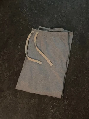Ljusgrå mjukisbyxor med dragsko - Säljer ett par ljusgrå mjukisbyxor med vita dragsnören och resår i midjan. Byxorna har sidofickor och är gjorda i mjukt bomullsmaterial, perfekta för chill dagar hemma eller när du vill vara bekväm.