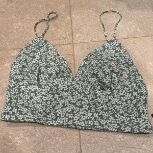 Grön blommig croptopp - Söt grön croptopp med vitt blommönster och tunna axelband från Lulus. Toppen har smockad rygg för extra komfort och bättre passform. Perfekt för varma dagar och enkel att matcha med både jeans och kjol. Aldrig använd, prislappen kvar. Säljs i befintligt skick. 