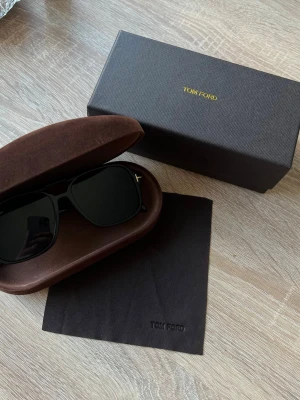 Svarta Tom Ford solglasögon - Snygga svarta solglasögon från Tom Ford med klassisk pilotform. Glasögonen har svarta bågar och mörka linser, samt den ikoniska T-detaljen vid gångjärnen. Kommer med originalfodral och putsduk.
