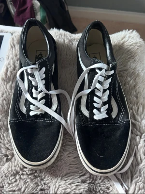 Svarta Vans Old Skool sneakers - Klassiska Vans Old Skool sneakers i svart canvas och läder med vit sula och vita snören. Skorna har den ikoniska vita sidoremsan och låg profil. Perfekta för en avslappnad streetstil.