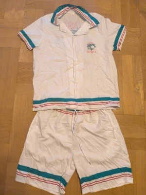 Casa Blanca vit med kanin - Vit pyjamas från Casa Blanca med kortärmad skjorta och shorts. Setet har turkosa och röda ränder på ärmar, krage, nederkant och benslut. Tryck med kanin och texten 'Casa Blanca Sport' på ryggen. Knappar framtill och dragsko i midjan. Perfekt för chillkvällar.