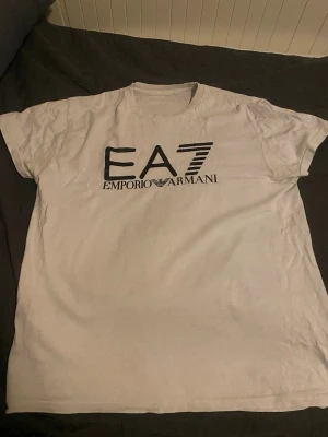 Vit EA7 Emporio Armani t-shirt - Snygg vit t-shirt från EA7 Emporio Armani med stor svart logga framtill. Klassisk rund halsringning och korta ärmar. Tillverkad i mjuk bomull som känns skön mot huden. Perfekt för dig som gillar stilrena och trendiga plagg.
