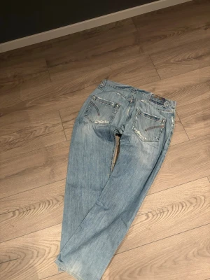 Ljusblå lite baggy dondup - Säljer ett par ljusblå jeans med raka ben och slitna detaljer framtill. Jeansen har fem fickor och dekorativa sömmar på bakfickorna. Perfekta för en avslappnad och trendig look.