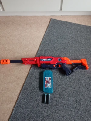 X-Shot leksaksgevär med magasin - Säljer ett X-Shot leksaksgevär i rött, orange och blått plastmaterial. Geväret har ett långt pipformat utförande och kommer med ett blått magasin. Perfekt för lek och actionfyllda strider med kompisar.