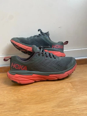 Hoka svarta sneakers med röd sula storlek 40&2/3 - Snygga sneakers från Hoka i svart mesh med ventilerande hål och en chunky röd sula. Skorna har klassisk låg profil, snörning och Hoka-logga på sidan. Perfekta för dig som gillar sportig och modern stil.