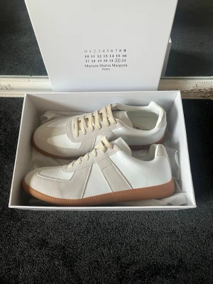 Maison Margiela vita sneakers - Maison Margiela sneakers i vitt läder och mocka med beige detaljer och gummisula. Klassisk låg modell med vita snören och stilren design. Perfekta för dig som gillar minimalistisk och trendig stil.