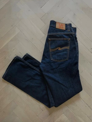 Mörkblå jeans från Nudie Jeans - Säljer ett par mörkblå jeans från Nudie Jeans med klassisk femficksmodell och raka ben. Jeansen har kontrastsömmar och en snygg läderpatch med logga bak i midjan. Materialet är kraftig denim i 100% bomull.