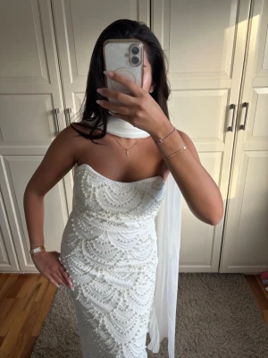 Vit långklänning med pärldetaljer - Elegant vit långklänning med pärlbroderier i vågmönster. Klänningen är axelbandslös och har en figurnära passform. Tillhörande vit sjal ger en extra touch. Perfekt för speciella tillfällen där du vill sticka ut. Endas använd i några få timmar🫶