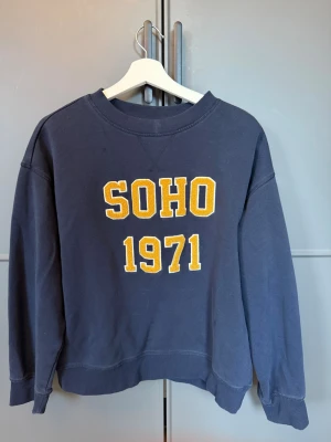 Mörkblå sweatshirt med SOHO 1971 - Snygg mörkblå sweatshirt med gul text 'SOHO 1971' på bröstet. Tröjan har rund halsringning, långa ärmar och ribbade muddar vid ärmslut och nederkant. Perfekt för en avslappnad och trendig look.