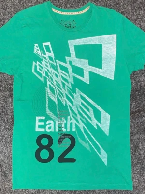 Grön t-shirt med grafiskt tryck - Snygg grön t-shirt från Discrete med ett stort grafiskt tryck i vitt och svart framtill. Trycket har texten 'Earth 82' och geometriska former. Perfekt för dig som gillar unika prints och vill sticka ut lite extra.