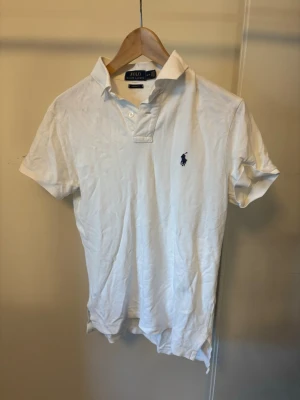 Vit pikétröja från Polo Ralph Lauren - Klassisk vit pikétröja från Polo Ralph Lauren med korta ärmar och blå logga broderad på bröstet. Tröjan har krage och knappar framtill, perfekt för en stilren och avslappnad look.