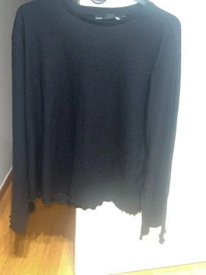 Svart långärmad tröja med vågig kant - Svart långärmad tröja i tunt, mjukt material med avslappnad passform. Tröjan har rund halsringning och rå, vågig kant nertill och vid ärmslut för en edgy look. Perfekt för dig som gillar minimalistisk stil med en twist.