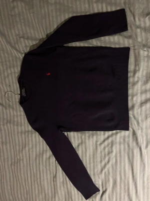Mörklila stickad tröja Polo Ralph Lauren - Snygg mörklila stickad tröja från Polo Ralph Lauren med klassisk rund halsringning och den ikoniska röda logotypen broderad på bröstet. Tröjan har långa ärmar och ribbade muddar vid ärmslut och nederkant. Perfekt för lager på lager under kalla dagar.