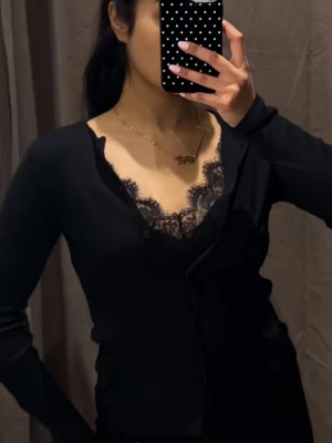 Svart cardigan med spets från Zara - Snygg svart cardigan från Zara med spetsdetaljer vid halsringningen. Långärmad och figurnära modell med knappar framtill. Perfekt för dig som gillar en stilren look med en touch av spets.