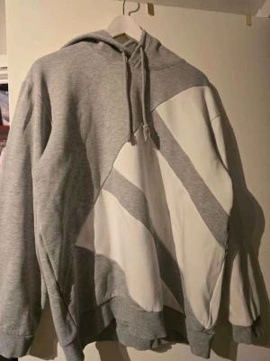 Grå/vit Adidas Original hoodie. Storlek L  - Säljer en grå hoodie med huva och dragsnören. Tröjan har en unik vit grafisk design med diagonala fält både fram och bak. Tillverkad i mjukt sweatshirtmaterial och har ribbade muddar vid ärmar och nederkant. Klassisk Adidas original hoodie. I nyskick. 