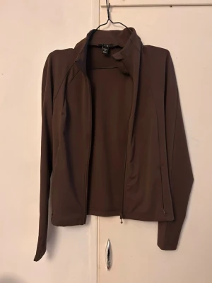 Brun zip-jacka med hög krage - Snygg brun jacka med dragkedja och hög krage. Jackan har långärmad design och är tillverkad i ett mjukt, stretchigt material som passar perfekt till vardags. Enkel och stilren look.