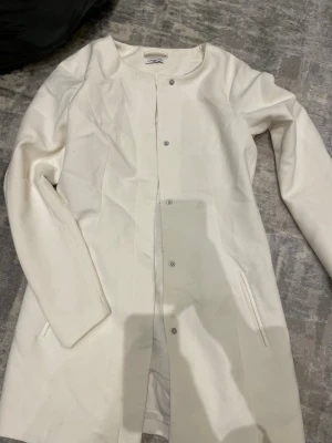 Vit rak kappa med tryckknappar - Stilren vit kappa med rak passform och tryckknappar framtill. Kappan har långa ärmar och diskreta fickor i sidorna. Materialet ser ut att vara mjukt och något tjockare, perfekt för kyligare dagar. Enkel och modern design som passar till det mesta.