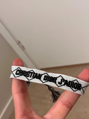 Christian Dior J'Adior armband - Snyggt armband från Christian Dior med texten 'J'Adior' i svart på vit bakgrund. Armbandet är vävt med tofsar i ändarna och har en trendig, ungdomlig stil. Perfekt accessoar för att lyfta din outfit.