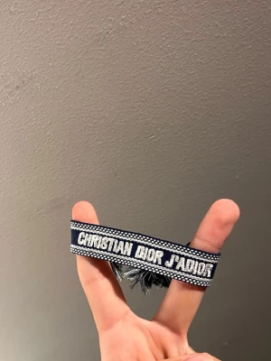 Blått Christian Dior J'Adior armband - Snyggt blått tygarmband från Christian Dior med texten 'J'Adior' broderat i vitt. Armbandet har justerbara band med tofsar i ändarna och en cool, avslappnad look. Perfekt accessoar för att lyfta din outfit med en exklusiv touch.