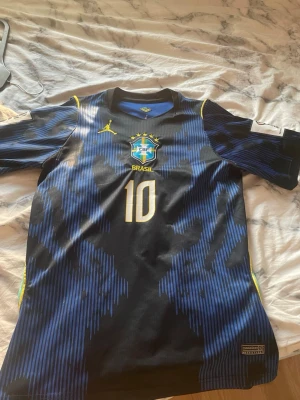 Brasilien Neymar Jr matchtröja blå - Säljer en blå och svart Brasilien matchtröja med Neymar Jr och nummer 10 på ryggen. Tröjan har gula detaljer, korta ärmar och officiella klubbmärket på bröstet. Perfekt för dig som älskar fotboll och vill stötta Brasilien.