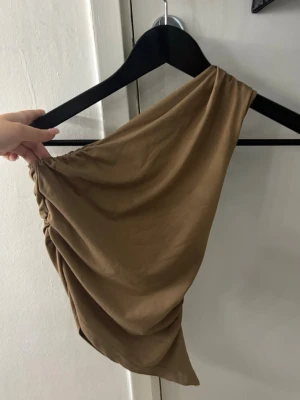 Brun oneshoulder topp - Snygg brun oneshoulder topp med draperad effekt. Toppen är ärmlös på ena sidan och har en asymmetrisk skärning som ger en trendig look.