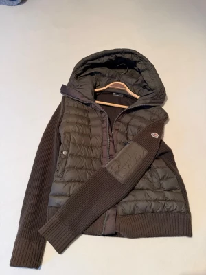 Oliv hybridjacka med huva - Snygg oliv hybridjacka med vadderad front och huva samt stickade ärmar och rygg. Jackan har dragkedja, två sidofickor och en patch på ärmen. Perfekt för lager-på-lager och chill höstväder.